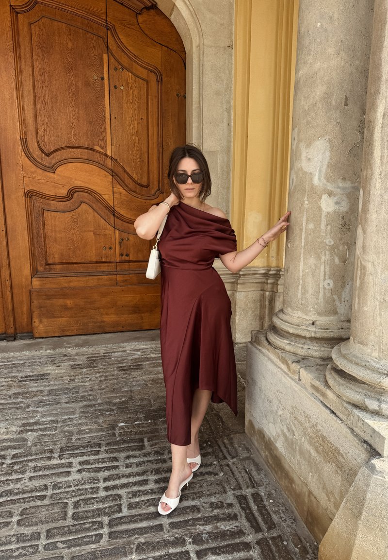 Γυναίκα με μπορντώ φόρεμα χωρίς ώμους και λευκά σανδάλια ποζάρει δίπλα σε πέτρινες κολόνες και μια μεγάλη διακοσμημένη ξύλινη πόρτα.