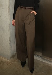 Personne portant un pantalon marron taille haute à jambes larges avec une ceinture, des chaussures noires à bouts pointus et un haut noir, debout près d'un mur en béton et d'une porte en métal.