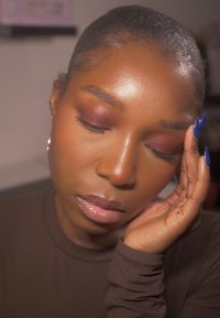 Nærbillede af en person med mørk hud og kort hår, iført en brun top. Karakteristika inkluderer dristige blå negle og blød mat øjenmakeup i burgundertoner.