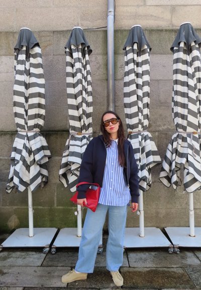 Mujer con gafas de sol y ropa casual está junto a cuatro paraguas cerrados a rayas negras y blancas contra una pared de piedra.