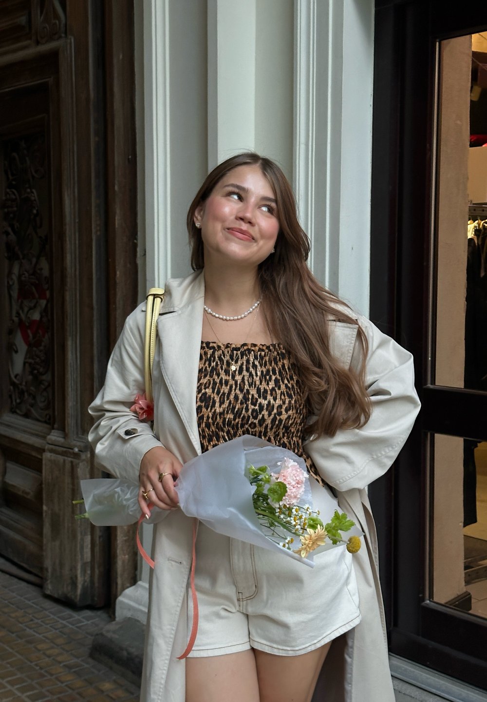 Beige trenchcoat over en leopardskinnetopp og hvite shorts. Holder en bukett pakket inn i papir, med rosa og gule blomster.