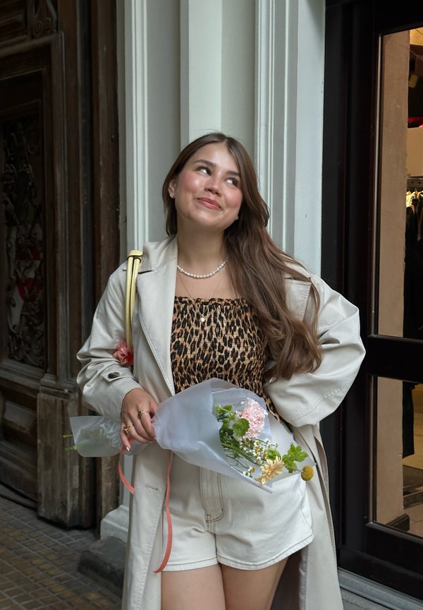 Beiger Trenchcoat über einem Leopardenmuster-Oberteil und weißen Shorts. Hält einen in Tissue eingewickelten Blumenstrauß mit pinken und gelben Blumen.