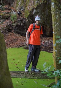 Oranje T-shirt met "TEKNO" erop gedrukt, zwarte broek en blauwe sportschoenen; staand op een boomstronk boven groen algwater in een bosachtige omgeving.