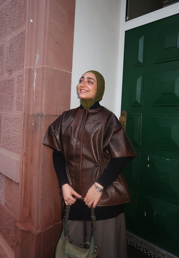Femme portant un hijab vert olive et une cape en cuir marron sourit en tenant un sac à main gris, debout près d'une porte verte et d'un mur en briques rouges.