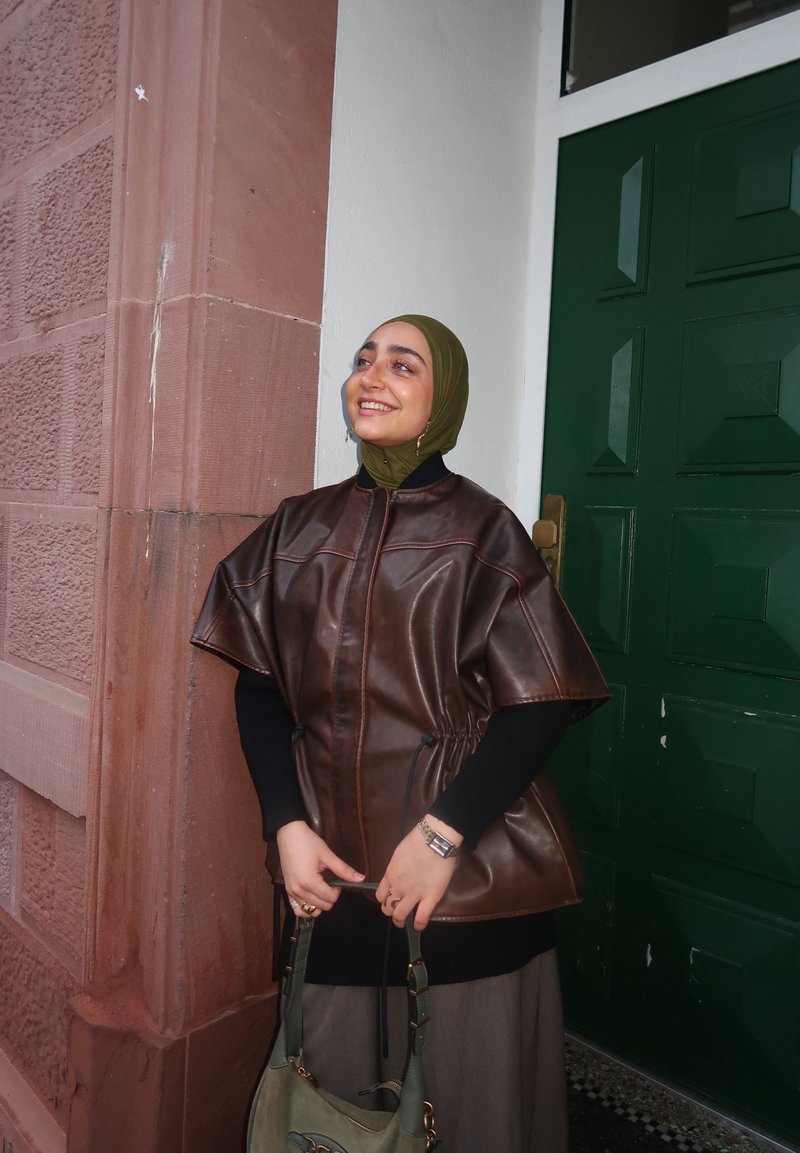 Donna con hijab verde oliva e mantella di pelle marrone sorride tenendo una borsa grigia, in piedi accanto a una porta verde e a un muro di mattoni rossi.