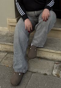 Γκρι φθαρμένα παντελόνια σε φαρδιά γραμμή, μαύρο μπουφάν με φερμουάρ και λευκές ρίγες, και καφέ ξύλινα υποδήματα με δύο λουράκια και λευκές λεπτομέρειες, καθισμένα σε σκαλιά.