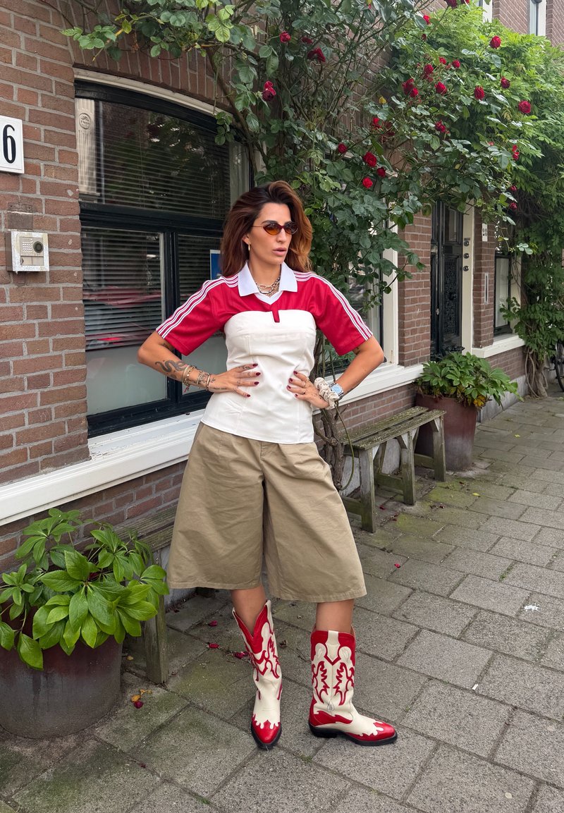 Woman wearing red and white top, beige wide-leg shorts, and red and white cowboy boots standing on a sidewalk near a brick building with rose bushes.