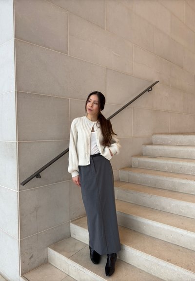 Cárdigan blanco de canalé sobre una blusa clara, combinado con una falda midi gris y botas negras hasta el tobillo, de pie junto a una escalera de piedra.