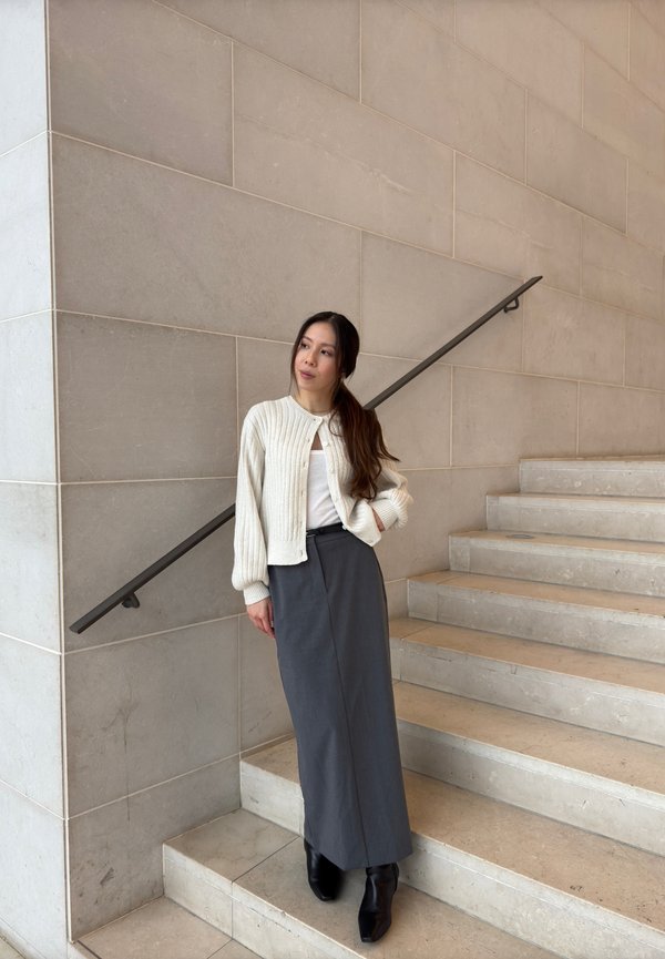 Cardigã branco canelado por cima de uma blusa clara, combinado com uma saia midi cinza e botas ankle pretas, de pé ao lado de uma escadaria de pedra.