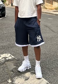 Ναυτικές αθλητικές βερμούδες με λευκές λεπτομέρειες και λογότυπο της New York Yankees, συνδυασμένες με λευκά αθλητικά παπούτσια Nike και λευκές κάλτσες.