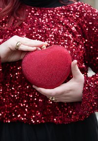 Personne portant un haut à sequins rouges tient une pochette rouge en forme de cœur ornée de petits cristaux rouges et d'un fermoir doré, montrant des bagues dorées aux doigts.