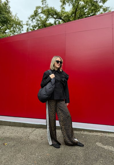 Blusa negra con detalles de volantes, pantalones anchos con estampado de leopardo y rayas blancas, y un bolso negro acolchado. Fondo rojo.