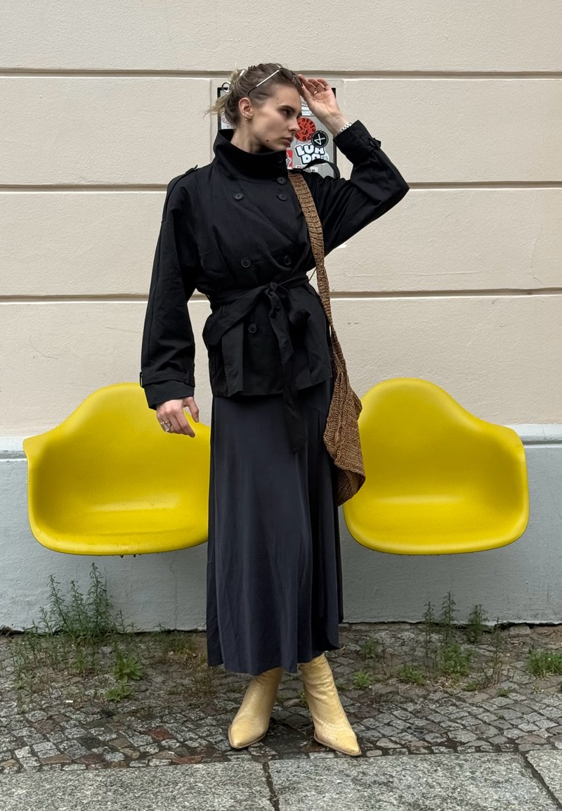 Femme en manteau noir et longue jupe ajuste ses lunettes, debout entre deux sièges en plastique moulé jaune fixés à un mur beige.