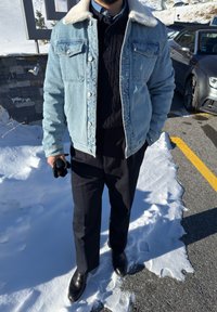 Veste en denim bleu clair avec un col en laine blanche, pull en maille torsadée noire, pantalon noir et chaussures en cuir noir. Sol enneigé visible.