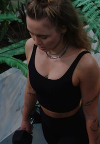 Must mustjas sportlik lühike top laiade rihmadega ja sobivad kõrge vöökohaga püksid. Silma paistvad tätoveeringud ja hõbedast kettidega kaelakeed. Hoidmas hantlit.