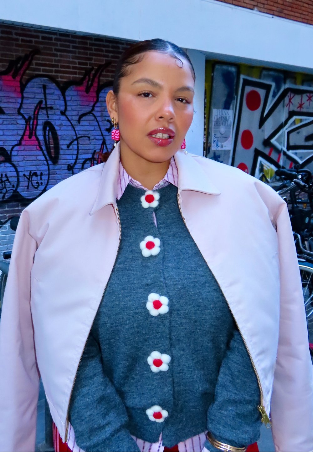 Rosa bomberjakke over en grå genser med blomsterknapper. Stripete skjorte under og rosa hengende øredobber. Urban graffiti-bakgrunn.