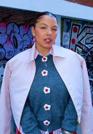 Chaqueta bomber rosa sobre un suéter gris con botones en forma de flores. Camisa a rayas debajo y pendientes colgantes rosas. Fondo de grafiti urbano.