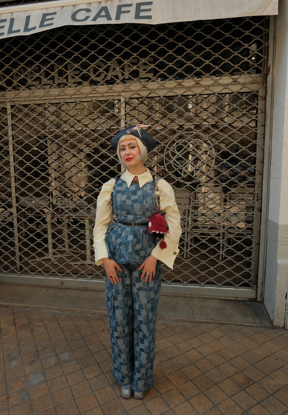 Persona con cabello corto rubio, vistiendo pantalones de cuadros azules y chaleco, blusa cremosa, sombrero azul con un lazo rosa, de pie frente a la puerta cerrada de un café.