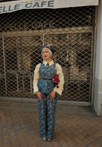 Persona con cabello corto rubio, vistiendo pantalones de cuadros azules y chaleco, blusa cremosa, sombrero azul con un lazo rosa, de pie frente a la puerta cerrada de un café.