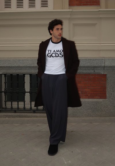 Abrigo negro sobre una camiseta gráfica blanca con el texto "TI AMO GCDS", combinado con pantalones oscuros holgados de color gris y zapatos negros sobre una superficie de adoquines.