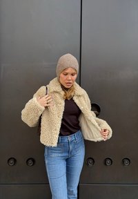 Beige fuskpälskappa med krage, långa ärmar och texturerad tyg. Bärs över en mörk topp och matchas med högmidjade blå jeans.