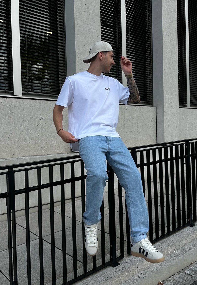 Vit bomulls-T-shirt med liten logotyp, ljusblå denimjeans och vita sneakers med svarta ränder, sittande på en svart räcke.