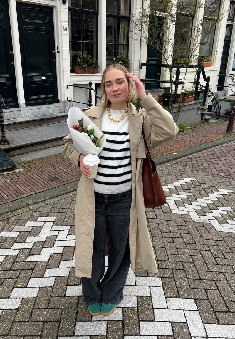Μπεζ παλτό τύπου trench πάνω από ένα ριγέ πουλόβερ, σκούρα τζιν και πράσινα παπούτσια. Κρατάει μια ανθοδέσμη με τουλίπες και ένα φλιτζάνι καφέ. Φόντο από τούβλινο δάπεδο.