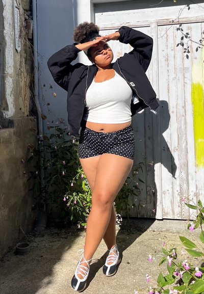 Mujer con pantalones cortos de lunares, camiseta blanca, chaqueta negra protegiéndose los ojos de la intensa luz solar al aire libre, cerca de una puerta de madera con flores.