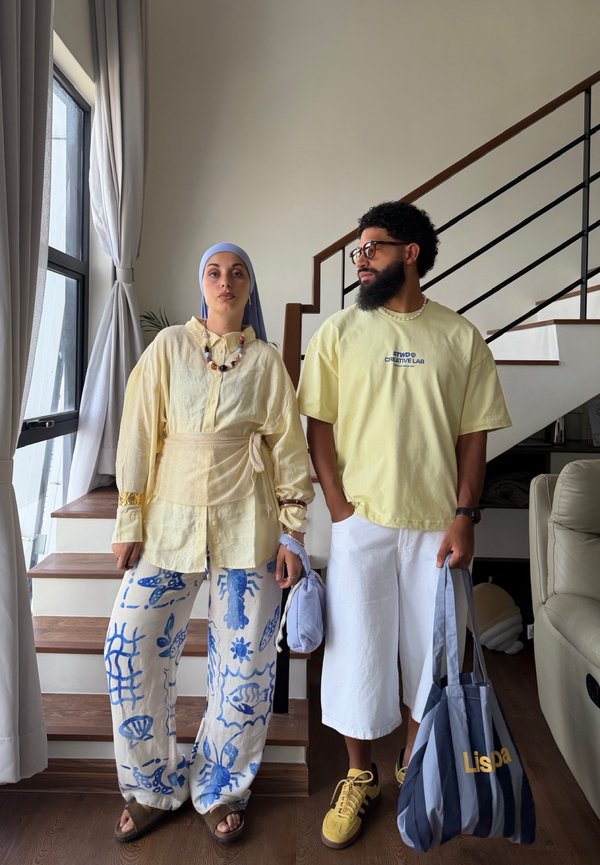 Zwei Personen stehen drinnen nahe einer Treppe; eine trägt gemusterte Hosen und ein Kopftuch, die andere weiße Shorts und hält eine gestreifte Tragetasche.
