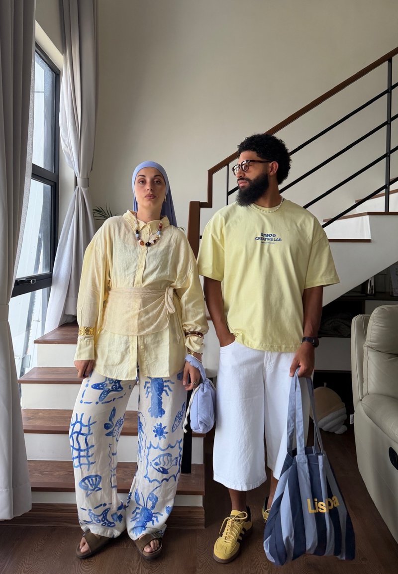 Deux personnes se tiennent à l'intérieur près d'un escalier ; l'une porte un pantalon à motifs et un foulard, l'autre porte un short blanc et tient un sac fourre-tout rayé.