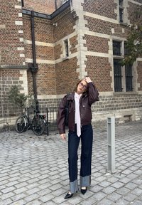 Brun läderpufferjacka, ljusrosa skjorta med slips, mörka jeans med fransiga fållar, svarta ballerinaskor. Bakgrund av kullersten och cykel.