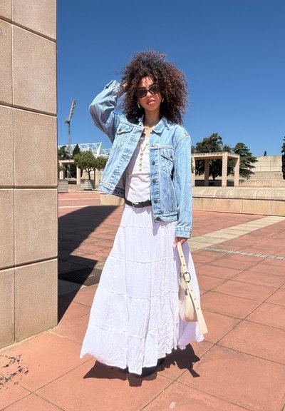 Chaqueta de denim sobre un vestido largo blanco de capas, complementada con un cinturón y un bolso ligero, en un ambiente soleado al aire libre.