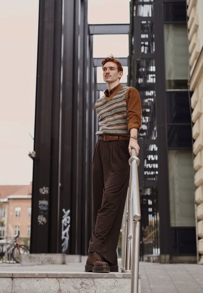 Chaleco de punto con rayas multicolores sobre una camiseta de manga larga marrón, combinado con pantalones de vestir marrones con pinzas y zapatos de cordones oscuros.