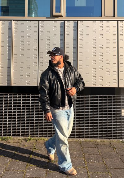 Hombre con chaqueta de cuero negra, vaqueros azul claro, gorra y gafas caminando por una calle pavimentada frente a una pared revestida de azulejos.