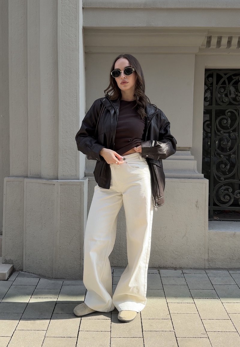 Femme portant une veste en cuir noire, un haut foncé, un pantalon blanc à jambes larges, des lunettes de soleil, debout devant une façade en pierre ornée.