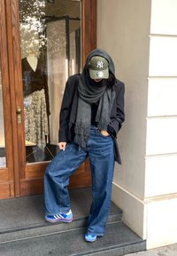 Bredde ben blå denim bukser, sort top, sort blazer, grå tørklæde, grøn baseball cap og blå sneakers med røde accenter, poseret på trin.
