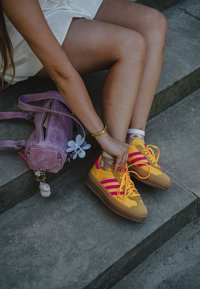 Gule joggesko med rosa striper og brune såler. En lilla teksturert veske med en blomsterdetalj hviler i nærheten. En person knytter skolissene.