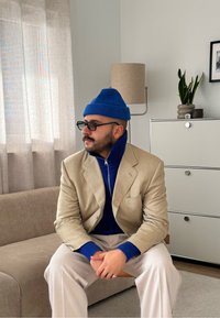 Blazer beige sur un col roulé bleu, associé à un pantalon clair. Un bonnet en maille bleu et des lunettes complètent le look. Cadre intérieur neutre.