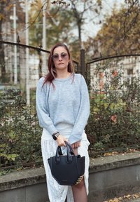 Zwarte handtas met gestructureerde vorm, voorzien van gouden studs, gecombineerd met een lichtblauwe geribbelde trui en een textuurrijke witte rok in de buitenlucht.