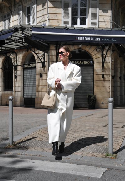 Abrigo oversized blanco con grandes solapas, llevado sobre botas negras. Bolsito beige con un asa corta. Gafas de sol oscuras. Entorno urbano con pavimento de piedra.