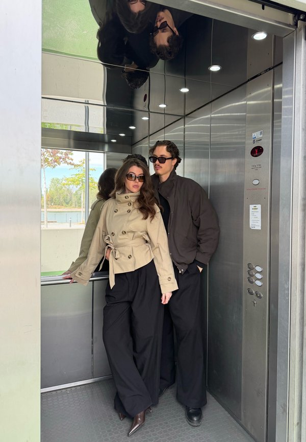 Une femme en veste beige et un homme vêtu de vêtements sombres posent dans un ascenseur, tous deux portant des lunettes de soleil.