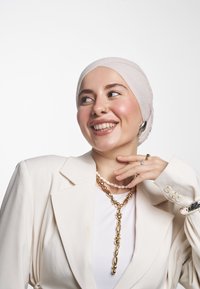 Vaalean beige bleiseri, jossa on jämäkät olkapäät, valkoisen paidan päällä, yhdistettynä kerrokselliseen kultaketju- ja helminauhaan. Silkkinen kangas ja kiiltävät yksityiskohdat.