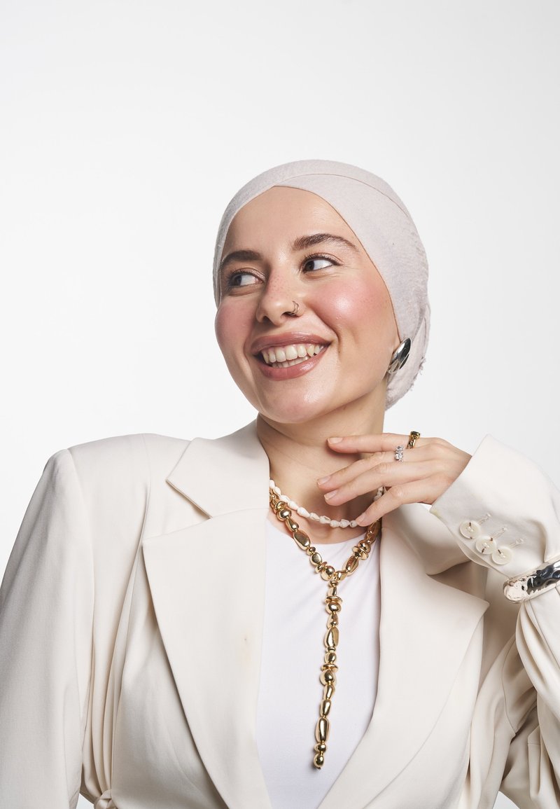 Vaalean beige bleiseri, jossa on jämäkät olkapäät, valkoisen paidan päällä, yhdistettynä kerrokselliseen kultaketju- ja helminauhaan. Silkkinen kangas ja kiiltävät yksityiskohdat.