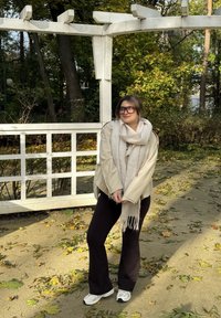 Beige-ylen suurikokoinen takki, mustat housut, valkoinen huivi ja valkoiset lenkkarit. Taustalla on puurakennelma ja pudonneita lehtiä.