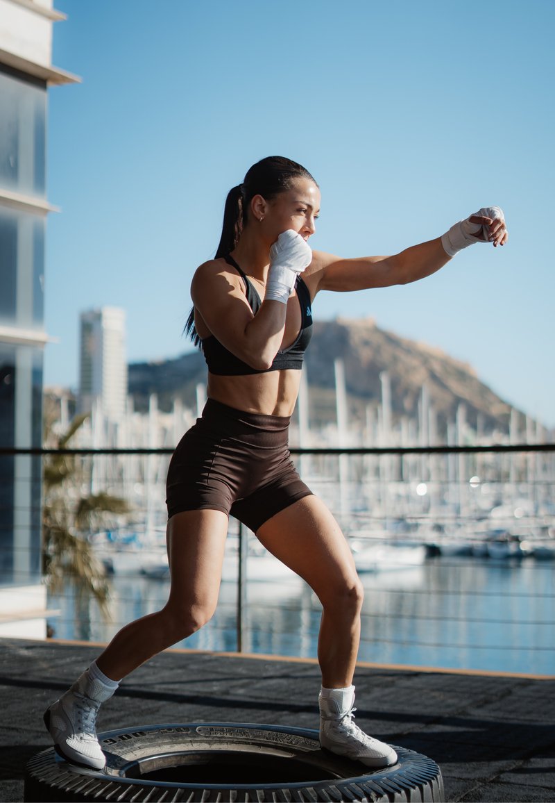 Γυναίκα με αθλητικά ρούχα και επιδέσμους στα χέρια στέκεται πάνω σε ένα ελαστικό έξω, ρίχνοντας μια γροθιά με μία μαρίνα και βουνά στο φόντο.