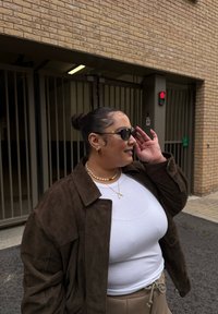 Brązowa zamszowa kurtka na białym prążkowanym topie na ramiączkach, beżowe spodnie ze ściągaczem, perłowy naszyjnik, okulary przeciwsłoneczne ze złoconymi detalami oraz kok z boku głowy.