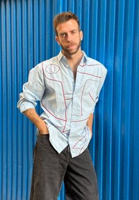 Lys blå button-up skjorte med røde geometriske mønstre, kombinert med mørke jeans. Avslappet passform med opprullede ermer, stående foran en blå bakgrunn.