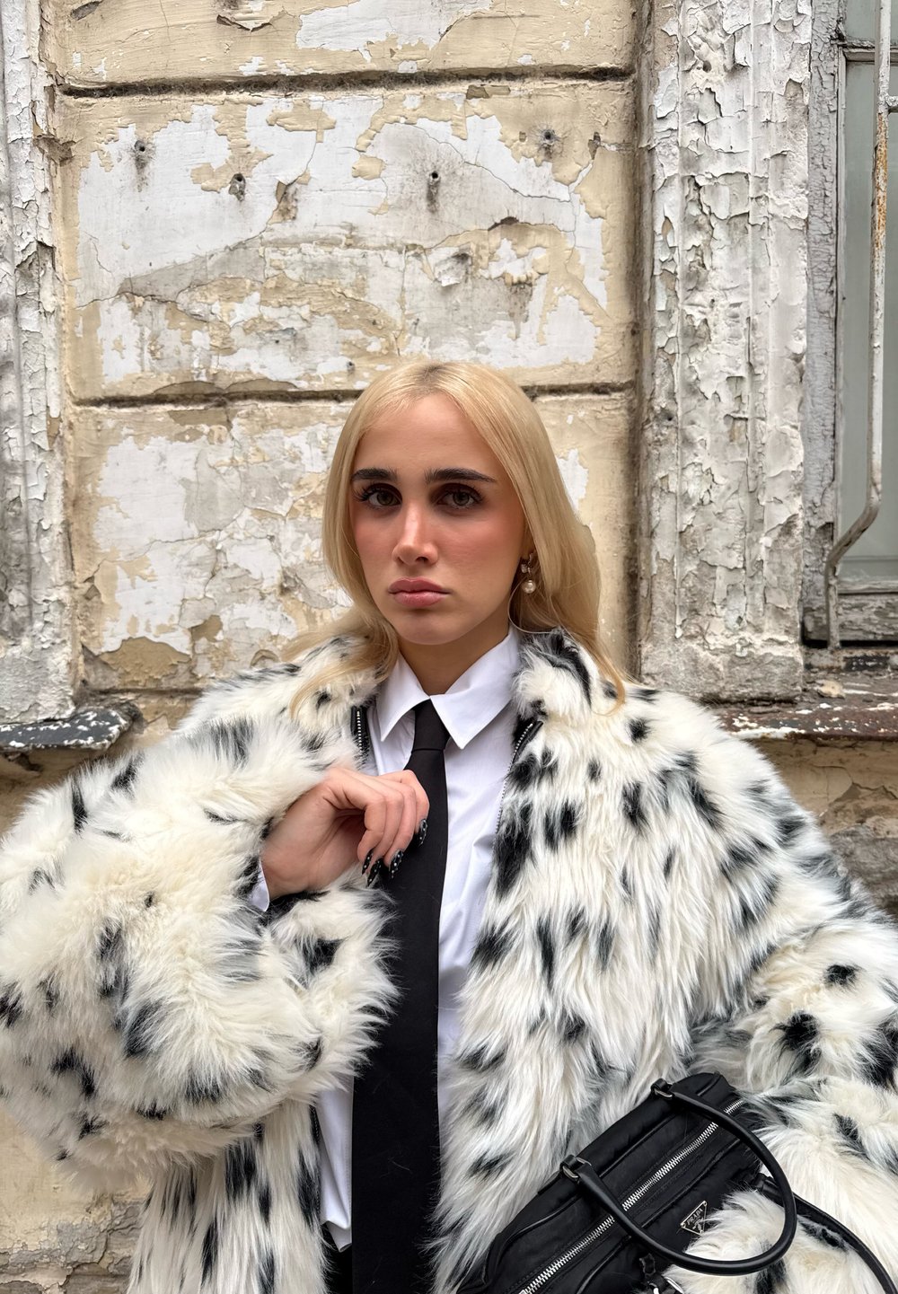 Femme blonde en cravate noire et chemise blanche porte un manteau en fourrure blanche surdimensionné avec des taches noires, tenant un sac à main noir contre un mur en plâtre écaillé.