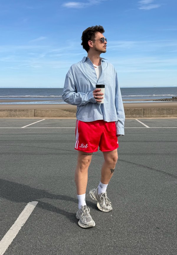 Mand i røde shorts og blå skjorte holder en kaffekop og står på en vej nær en strand med roligt hav og klar himmel.