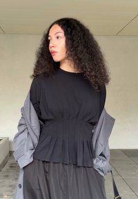 Top negro con pliegues y mangas cortas, combinado con un abrigo trinchera gris claro. Textura suave, escote redondo, llevado sobre una falda fluida.